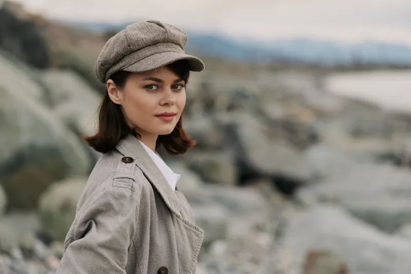 Young woman in vintage plaid coat and cap stands outdoors near rocky shore, looking at camera with soft smile. Moody cloudy sky and blurred sea background create calm atmosphere.