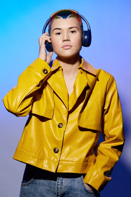 Young woman with colorful hair wearing a yellow shiny jacket and headphones against a blue-purple gradient background, expressing calm and focus in a modern style.