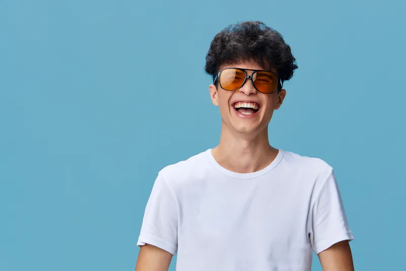 Smiling man wearing orange sunglasses and white t-shirt laughing against blue background with curly hair and joyful expression.