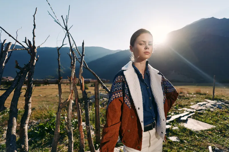 Woman portrait wearing a shearling jacket in mountains countryside at sunrise with sun near rustic fence, fashion model posing outdoors, relaxed confident expression and scenic landscape.