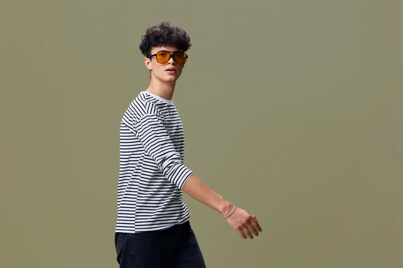 Young man with striped shirt and sunglasses walking confidently against neutral background in casual style, showcasing modern fashion and cool attitude.
