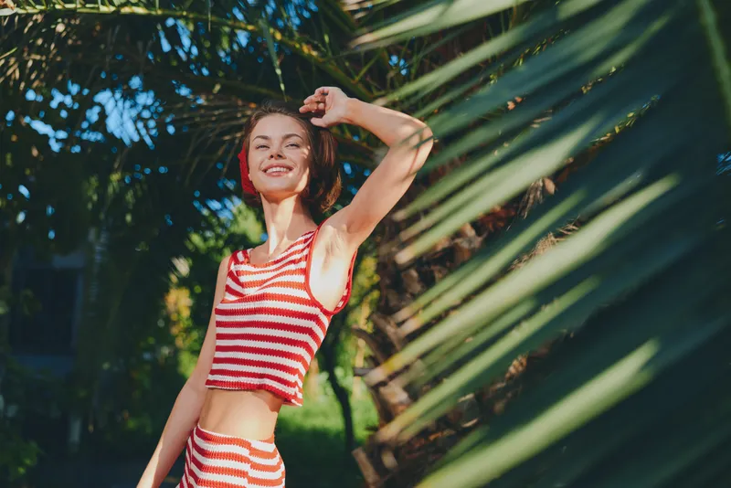 A cheerful woman in a red and white striped swimsuit stands outdoors among greenery, smiling brightly in warm sunlight, exuding carefree summer energy and relaxed confidence.
