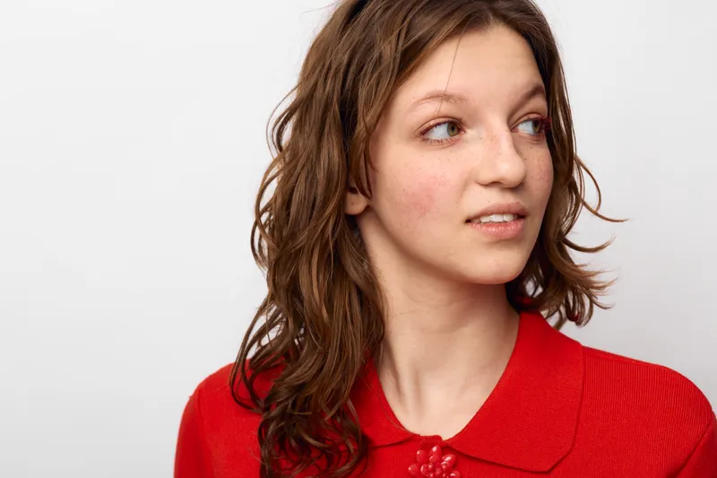 Young woman with natural skin and brown hair looks sideways wearing a red top. Studio portrait on white background. Concept of natural beauty and casual style.