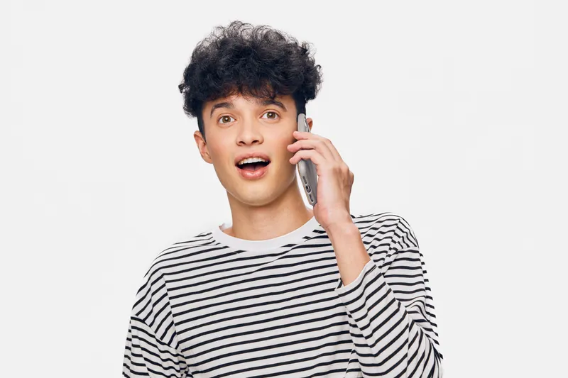 young man talking on phone with surprised expression in casual striped shirt against white background, communication, technology, positive emotion