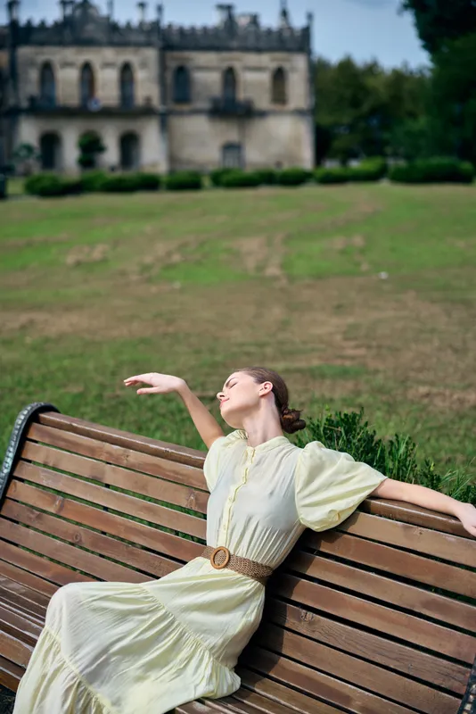 Vintage dress clad woman lounges on a wooden bench in a sunlit park, posing gracefully with one arm extended, a historic building visible in the distance.