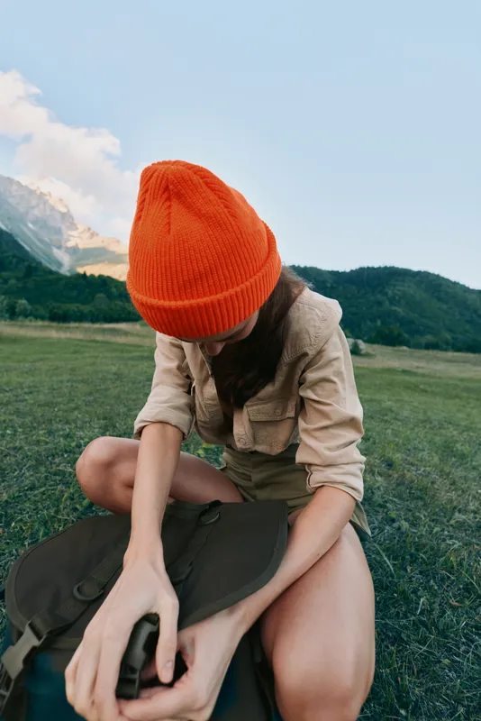Woman with orange beanie sits in a grassy meadow, organizing a backpack, mountains in the background, casual outdoor lifestyle, serene moment in nature and fresh air.