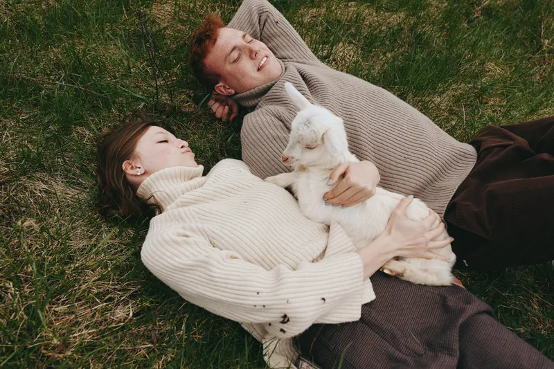 A relaxed couple lies on the grass, embracing a small goat kid as sunlight warms a calm yard, capturing lifestyle moments, affection, and bond between humans and animal companion.