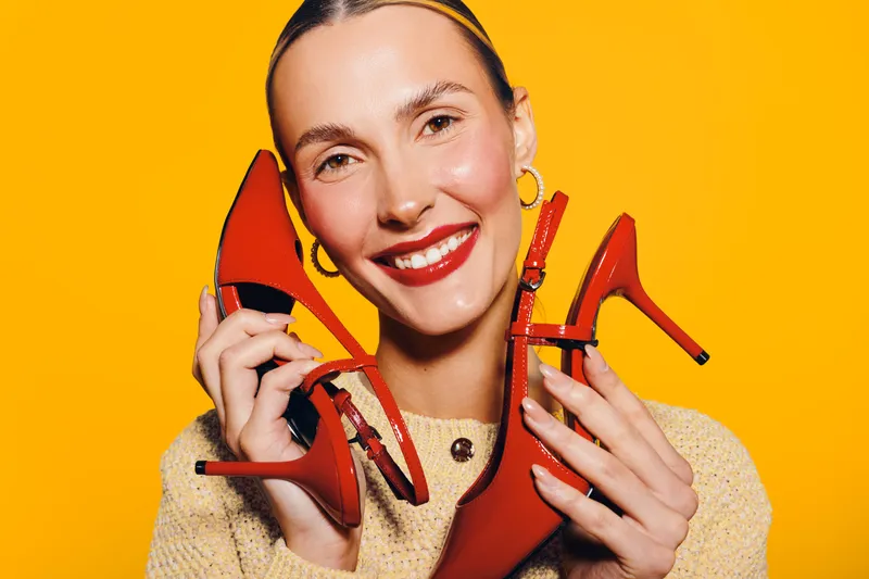 Smiling woman holds red high heel shoes near face on bright yellow background. Concept of fashion, style, footwear, and happiness with vibrant studio lighting and cheerful mood.
