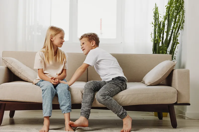 Two children sitting on beige sofa indoors smiling and talking, casual clothes, bright room with natural light and green plant in background.