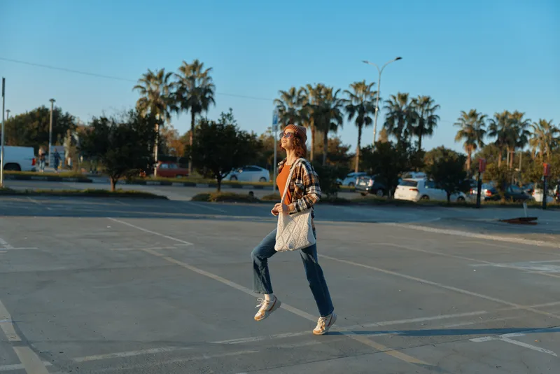 Woman jumping in an urban parking lot with a canvas tote, palms in background and warm golden light, candid lifestyle portrait highlighting authenticity, mindful living and emotional storytelling.