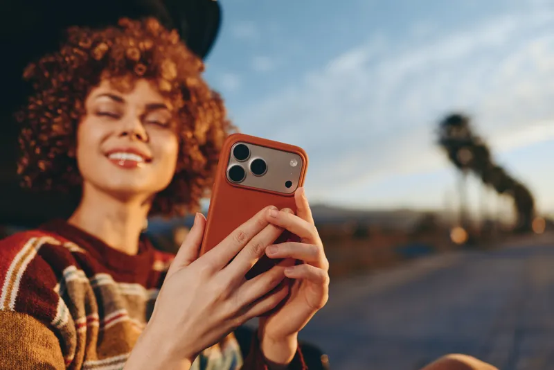 Smiling woman lifestyle outdoors at dusk, curly hair, holding smartphone with brown case, taking selfie with both hands, relaxed portrait near seaside horizon, warm sunset light, soft glow