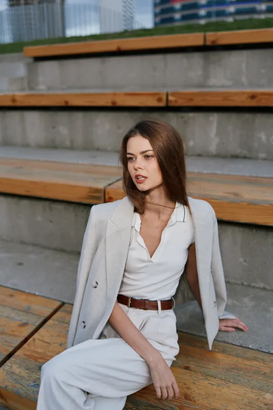 Businesswoman in elegant attire sitting on wooden steps, showcasing confidence and professionalism in a modern urban setting with a stylish outfit.