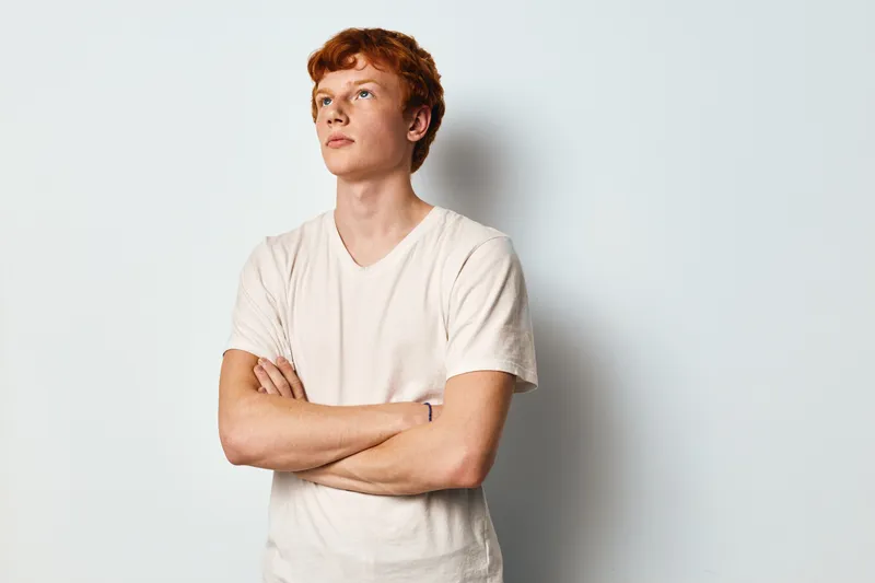Young man with red hair and crossed arms standing against white background looking up thoughtfully. Casual male wearing white t shirt in studio for portrait or lifestyle.