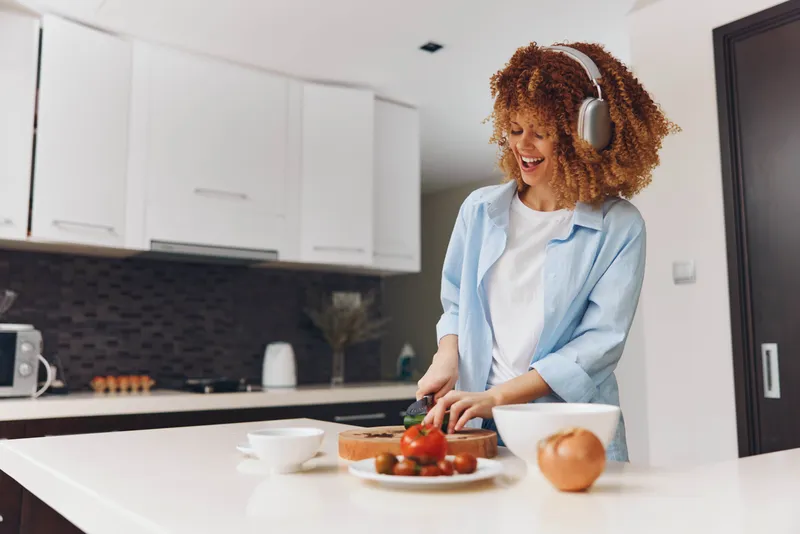 Женщина в наушниках готовит полезный перекус на кухне, в фокусе ломтики хлеба и помидор