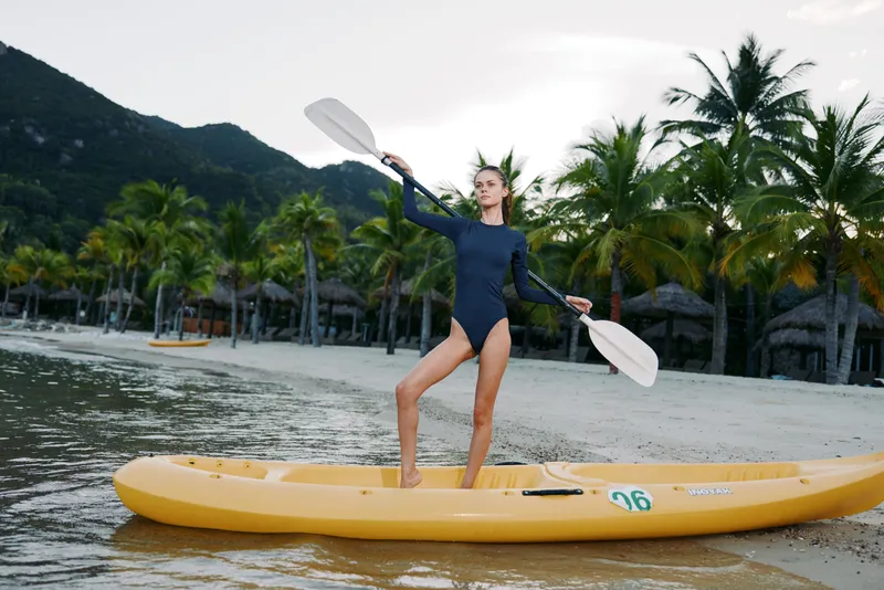 Dynamic scene features a kayaker balancing on a bright yellow kayak, paddle raised high while gliding over calm shoreline waters beneath a clear sky.