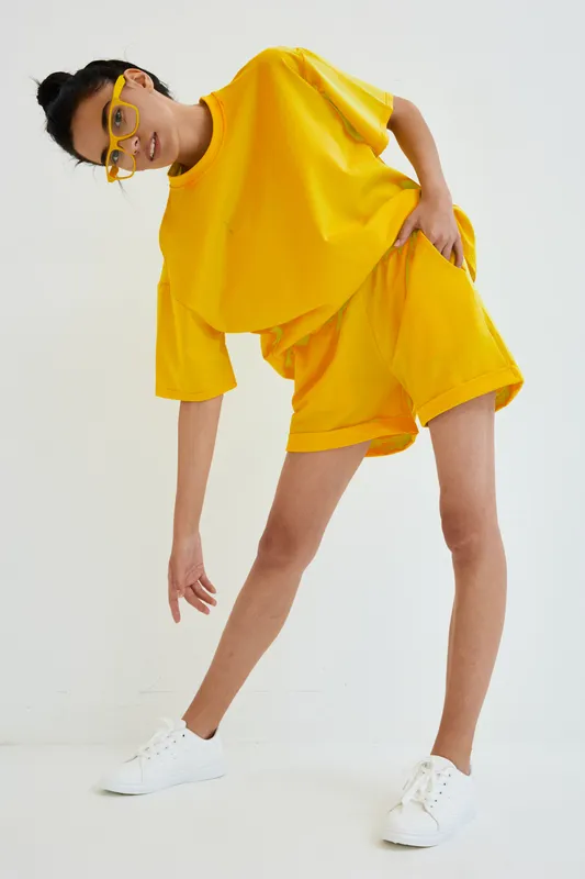 Young Asian woman in bright yellow oversized outfit and matching glasses posing against white background, showcasing modern casual style and playful attitude.