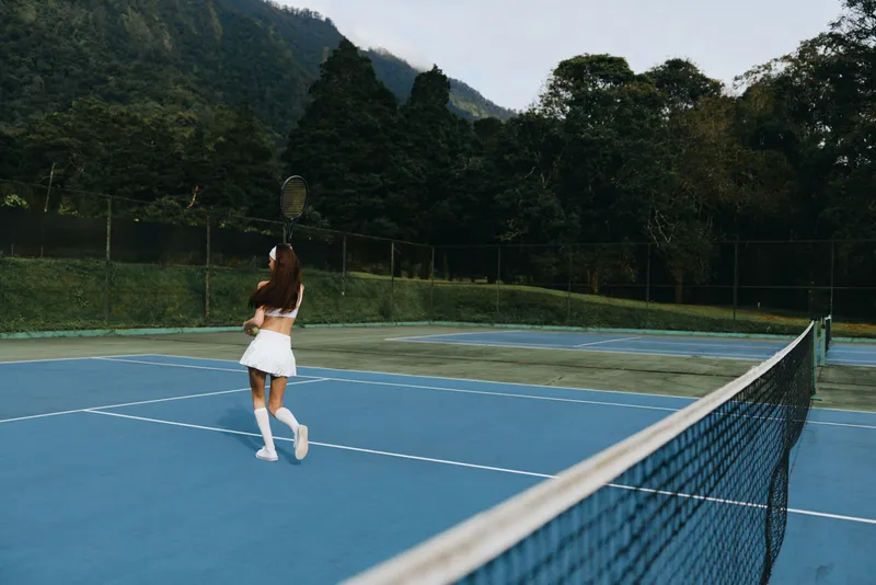Athletic young woman in white tennis outfit practicing on a blue court surrounded by lush green trees, showcasing focus and determination in a serene setting