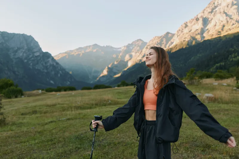 A woman stands in a meadow with towering mountains in the distance, arms outstretched, soaking in fresh air and freedom during an outdoor hiking experience.
