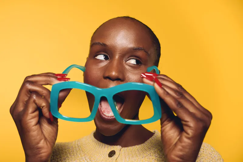 Dark skinned African woman with short hair holding oversized blue glasses near her mouth, looking sideways on yellow background. Concept for fun, fashion, and playful mood.