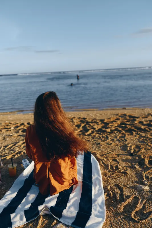 Young woman with long brown hair sits on a striped towel on the sandy beach watching the calm ocean during a peaceful late afternoon.