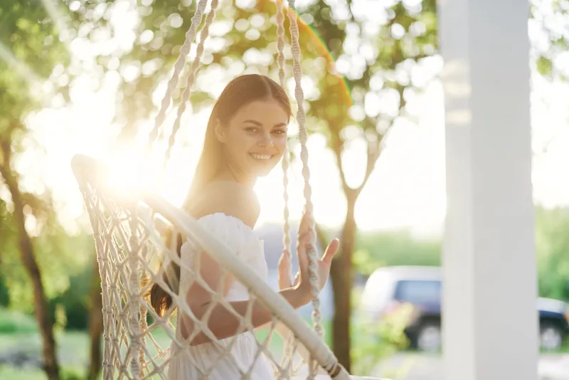 Young woman smiling in a white off shoulder dress on a swing, surrounded by greenery and sunlight, conveying joy and relaxation in a summer setting