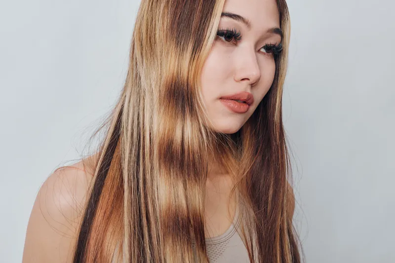 Young woman with long straight hair looks sideways, wearing casual tank top. Beauty style portrait with natural makeup and neutral expression on plain background.