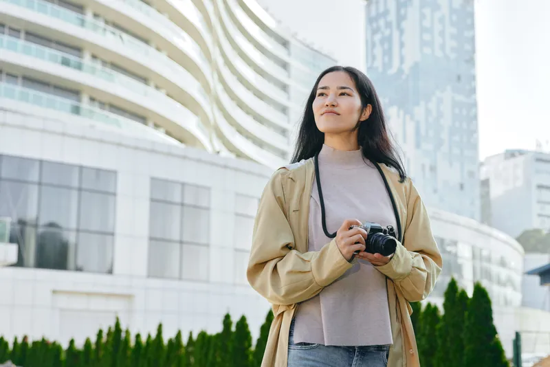 Woman with camera in hand stands outdoors amid modern buildings, exploring urban scenery and street photography, showcasing curiosity, travel vibes, and creative exploration in a city environment.