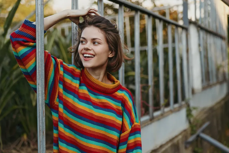 woman smiling wearing a colorful sweater portrait in an outdoor casual setting, joyful young model leaning on greenhouse frame, soft light, cozy autumn fashion candid mood