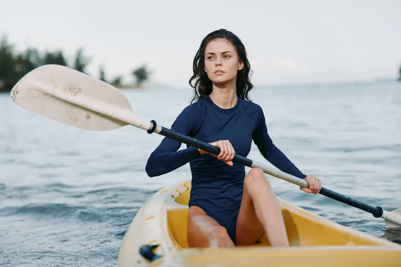 A focused young woman paddles a yellow kayak across calm water, wearing a dark long sleeve top and shorts, capturing a dynamic outdoor sport moment.