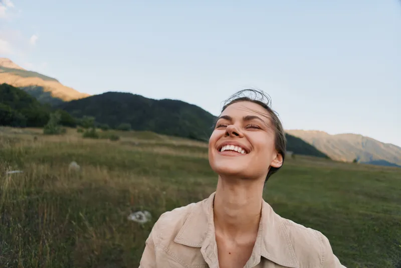 Smiling woman with closed eyes enjoying nature outdoors in a field with mountains in the background. Concept of happiness, lifestyle, and freedom in natural light.