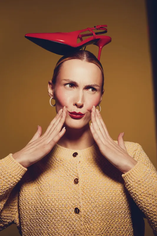Woman with red high heel shoe on head touches cheeks with hands, wearing yellow sweater and gold earrings. Concept for fashion, style, and quirky portrait with warm lighting.
