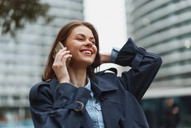 Young woman in a trenchcoat chats on a phone in a cityscape beside a skyscraper, smiling with confidence amid urban bustle, denim shirt peeking under the coat as sunlight highlights the street scene
