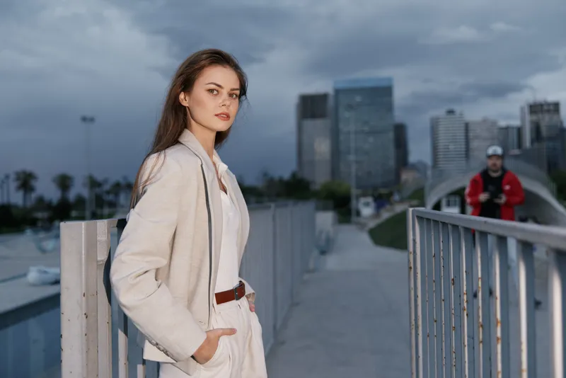 Confident woman in neutral suit posing against urban cityscape background during overcast day, fashion style with modern city and moody sky
