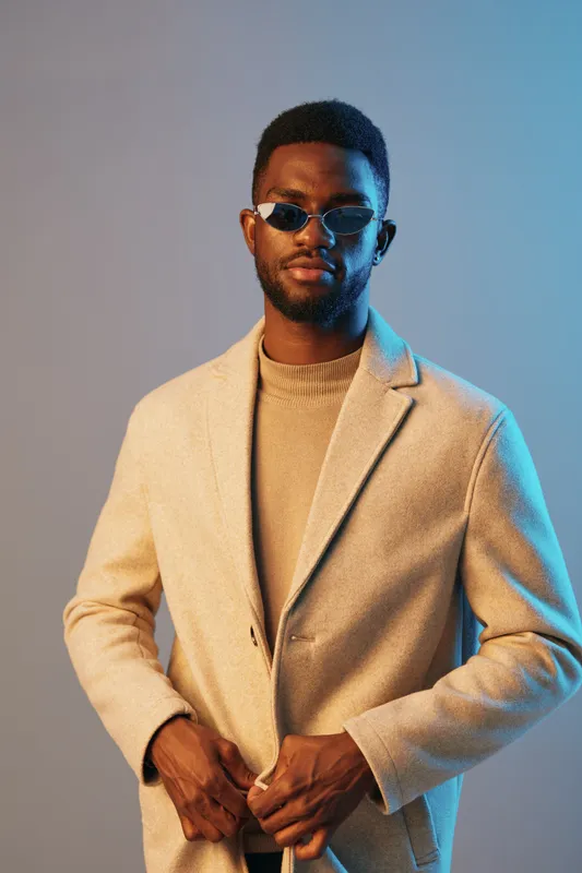 Stylish young Black man posing confidently in a beige coat and sunglasses against a blue background, conveying a modern and chic fashion concept