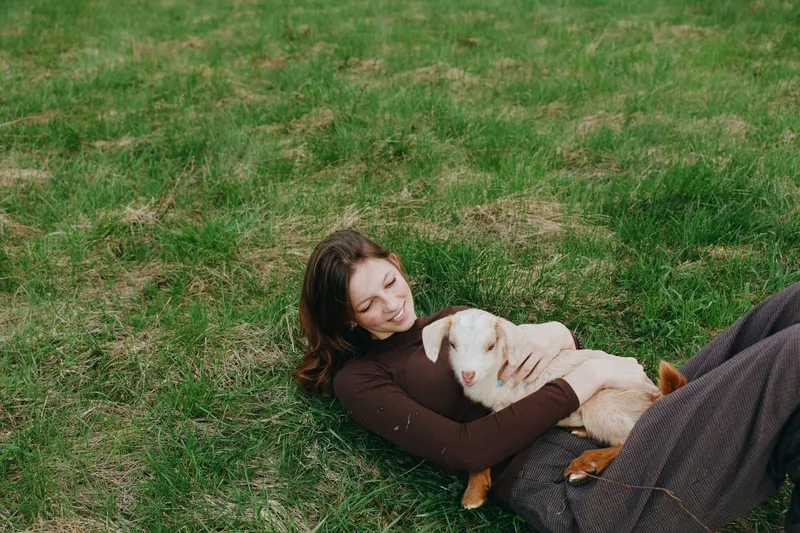 A relaxed woman lies on the grass, cuddling a gentle goat kid in a sunny outdoor setting. A candid lifestyle moment that conveys calm, companionship, and connection with animals.