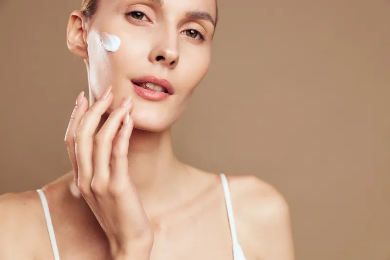 Young woman applies facial cream on cheek with hand, shows natural skin care routine, looks serene and fresh, isolated on beige background, wearing white top, beauty concept.