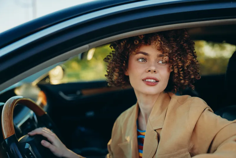 Smiling woman with curly hair sits behind the wheel in a car, wearing a beige jacket. Lifestyle moment captured with warm retro film color, emphasizing casual driving and everyday comfort.
