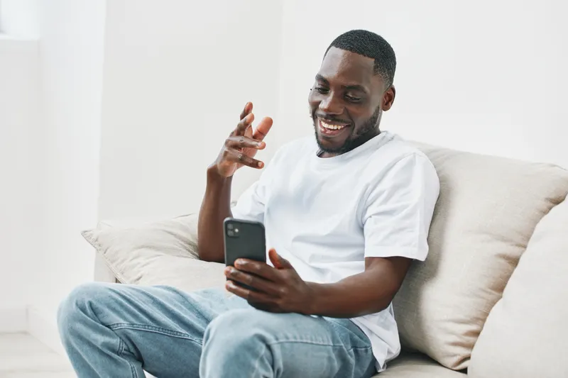 Smiling young African American man enjoying a video call on his smartphone while sitting on a cozy sofa in a bright living room, showcasing modern casual style