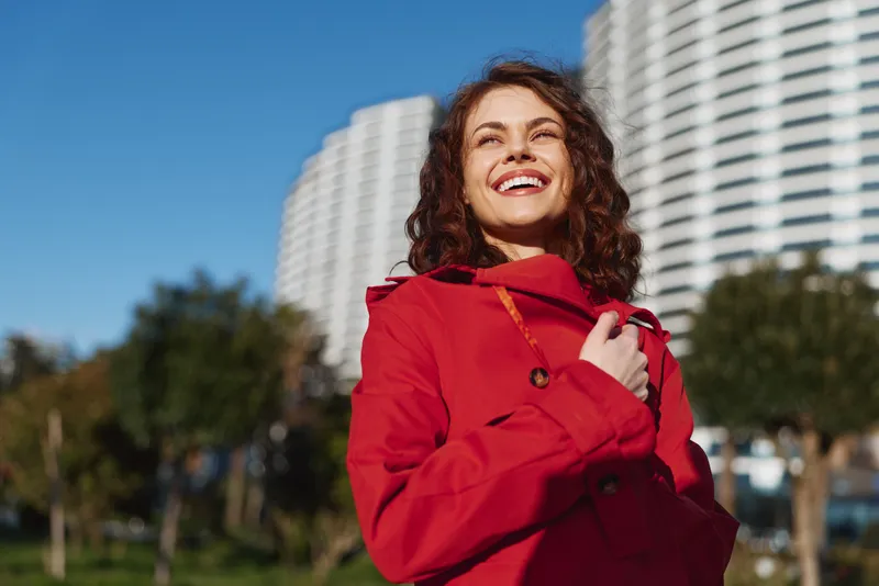 A happy young woman in a red coat smiles outdoors with sunlight on her face, with modern buildings and trees in the background on a clear day.