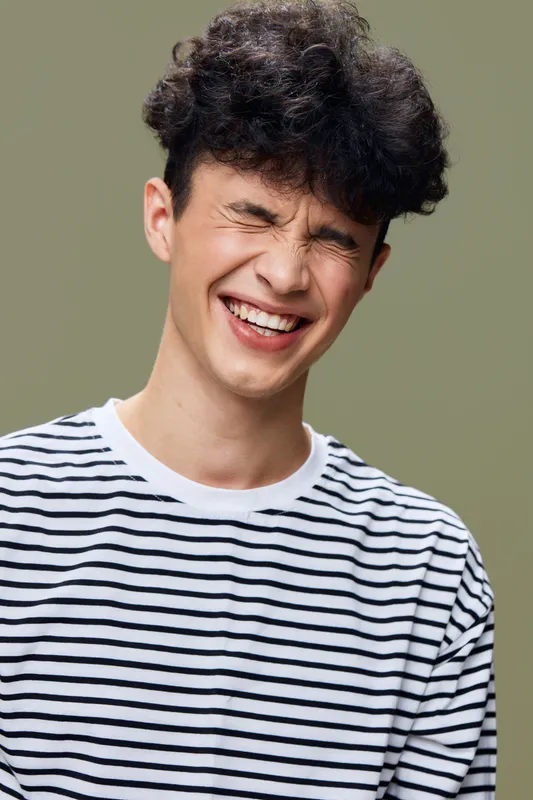 smiling young man in striped shirt with curly hair enjoying joyful moment, eyes closed, beige background, natural expression, casual style.