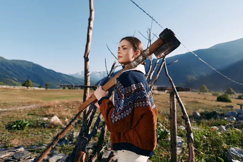 Woman axe mountains rural sweater field wood outdoors, woman with axe over shoulder in mountain field wearing patterned sweater near wooden fence in warm sunlight preparing firewood.