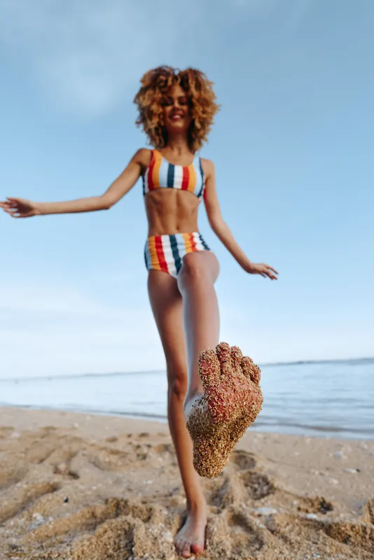Young woman with curly hair wearing a striped swimsuit joyfully kicks sand on a sunny beach near calm water, expressing carefree summer fun and relaxation.