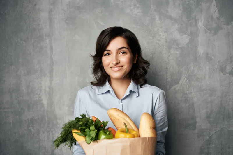 Улыбающаяся женщина с пакетом продуктов, включая овощи, совершает покупки в супермаркете