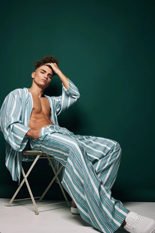 Young man wearing striped pajamas sitting on chair with hand in pocket, casual relaxed pose and fingers through hair in front of green background with cool fashion attitude