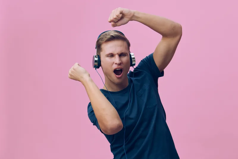 Joyful young man wearing headphones enjoying music, raising his arm in celebration against pink background, casual clothing, male, expression of happiness, lifestyle concept, studio shot, color image.