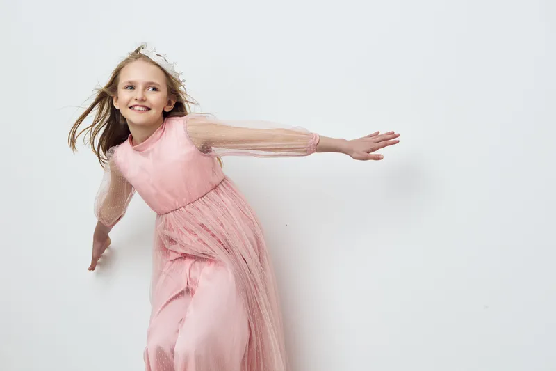 Delightful girl in a flowing pink dress, joyfully dancing against a minimalist background, capturing pure happiness and innocence in her playful expression