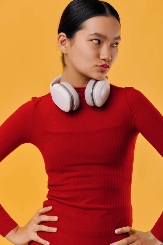 Young woman in red sweater and white headphones stands with hands on hips against orange background showing confident expression. Casual style and technology accessory in portrait.