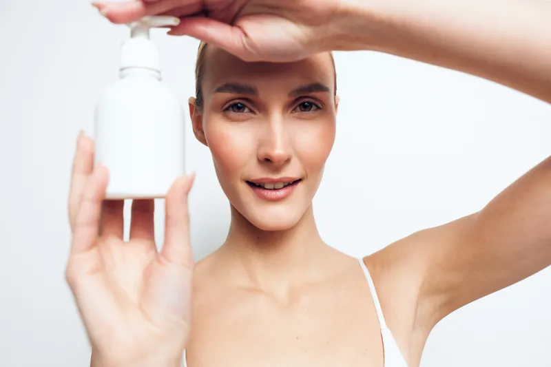 Young woman with clear skin holding white cosmetic bottle close to face, natural makeup, smiling gently, bright studio background, skincare concept, fresh beauty routine.