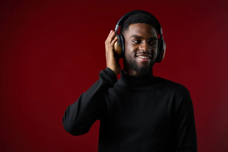 Young Black man in a black turtleneck enjoying music with headphones against a vibrant red background, embodying joy and passion for music