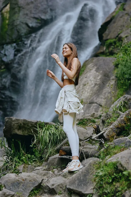young woman hiking near a waterfall in nature wearing sporty clothes, enjoying outdoor adventure and fresh air with green cliffs around